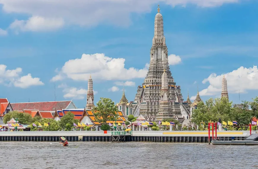 Vista do Templo Wat Arun às margens do Rio Chao Phraya, em Bangkok, com sua torre central ornamentada destacando-se contra um céu azul com nuvens, rodeado por edifícios coloridos e bandeiras tailandesas.