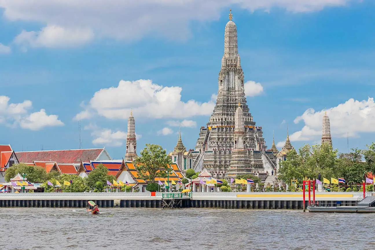 Vista do Templo Wat Arun às margens do Rio Chao Phraya, em Bangkok, com sua torre central ornamentada destacando-se contra um céu azul com nuvens, rodeado por edifícios coloridos e bandeiras tailandesas.