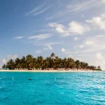 Ilha tropical cercada por mar azul-turquesa, com praia de areia branca, coqueiros e guarda-sóis coloridos sob um céu azul com nuvens claras.