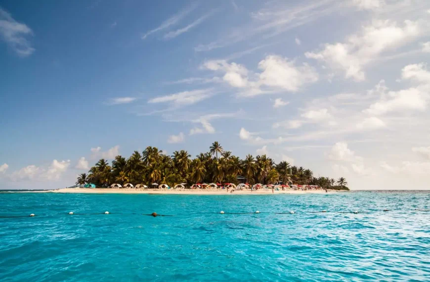 Ilha tropical cercada por mar azul-turquesa, com praia de areia branca, coqueiros e guarda-sóis coloridos sob um céu azul com nuvens claras.