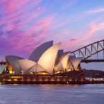 Vista do Sydney Opera House ao entardecer, com suas conchas iluminadas refletindo na água do porto, a Harbour Bridge ao fundo e o céu em tons de rosa e azul.