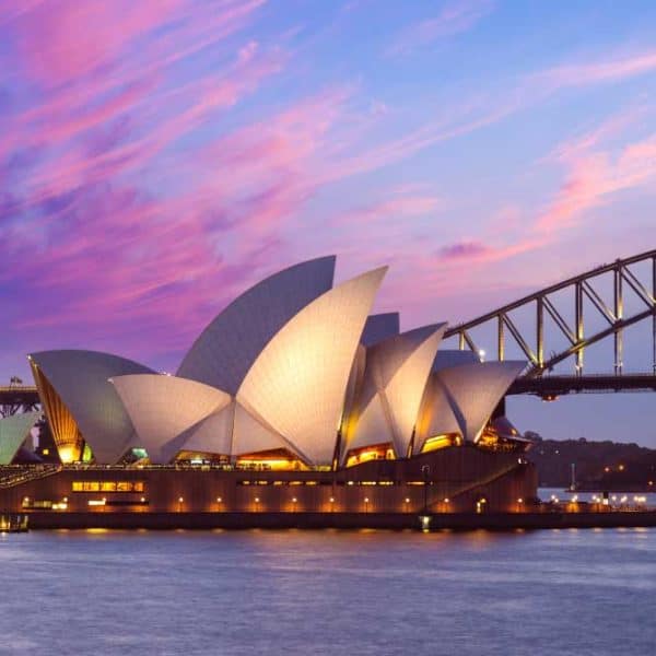 Vista do Sydney Opera House ao entardecer, com suas conchas iluminadas refletindo na água do porto, a Harbour Bridge ao fundo e o céu em tons de rosa e azul.