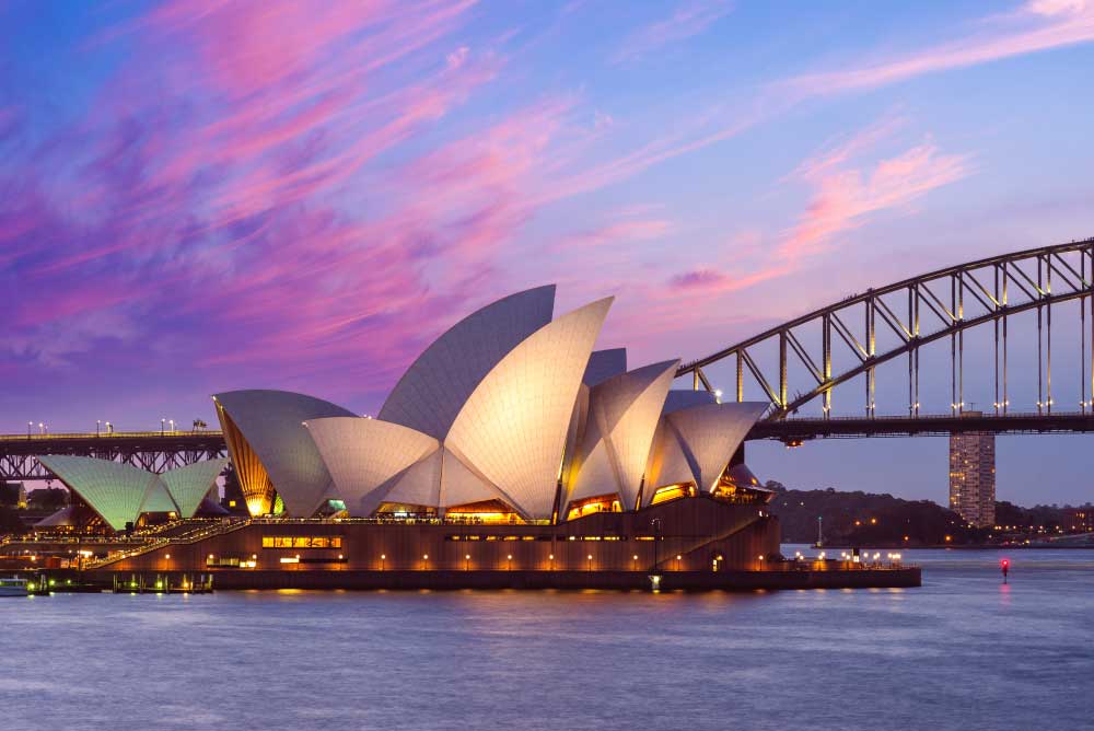 Vista do Sydney Opera House ao entardecer, com suas conchas iluminadas refletindo na água do porto, a Harbour Bridge ao fundo e o céu em tons de rosa e azul.