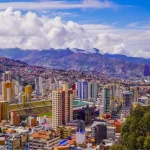 Vista panorâmica de uma cidade grande situada em um vale montanhoso, com muitos prédios altos e coloridos espalhados entre encostas íngremes. No centro, há um estádio de futebol de grande porte. Ao fundo, montanhas elevadas e nuvens densas completam a paisagem.