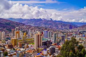 Vista panorâmica de uma cidade grande situada em um vale montanhoso, com muitos prédios altos e coloridos espalhados entre encostas íngremes. No centro, há um estádio de futebol de grande porte. Ao fundo, montanhas elevadas e nuvens densas completam a paisagem.
