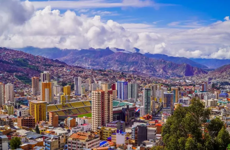 Vista panorâmica de uma cidade grande situada em um vale montanhoso, com muitos prédios altos e coloridos espalhados entre encostas íngremes. No centro, há um estádio de futebol de grande porte. Ao fundo, montanhas elevadas e nuvens densas completam a paisagem.