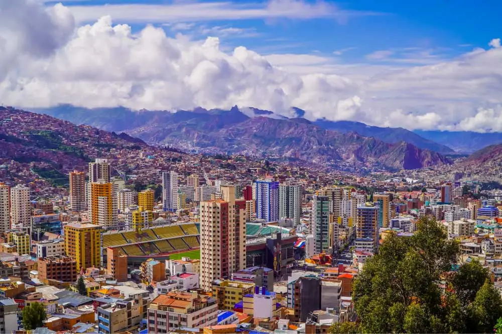 Vista panorâmica de uma cidade grande situada em um vale montanhoso, com muitos prédios altos e coloridos espalhados entre encostas íngremes. No centro, há um estádio de futebol de grande porte. Ao fundo, montanhas elevadas e nuvens densas completam a paisagem.