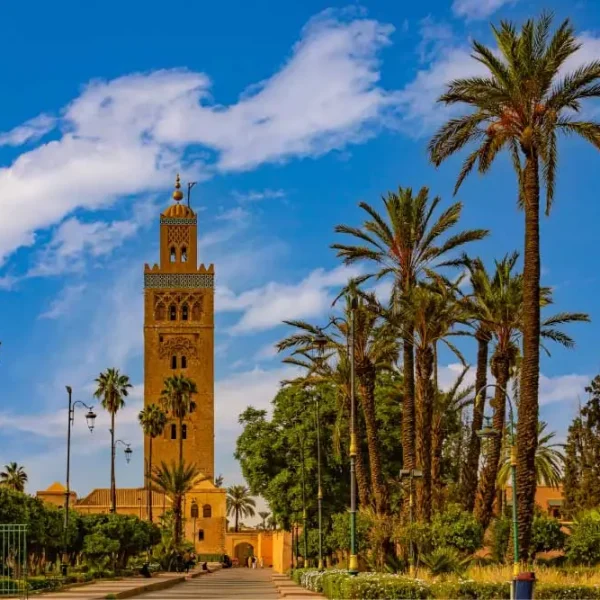 Avenida arborizada com palmeiras levando a um alto minarete de mesquita em tons de areia, sob céu azul com nuvens leves, em um ambiente urbano ensolarado