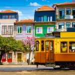 Um bonde elétrico histórico amarelo e marrom passando em frente a uma fileira de edifícios coloridos em Porto, Portugal, com árvores frondosas à esquerda sob um céu azul.