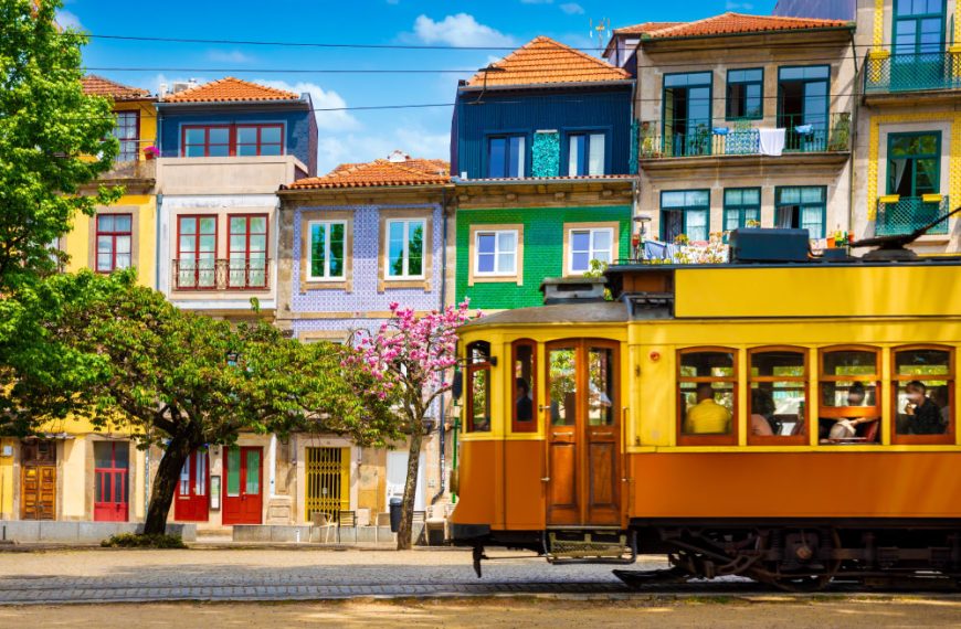 Um bonde elétrico histórico amarelo e marrom passando em frente a uma fileira de edifícios coloridos em Porto, Portugal, com árvores frondosas à esquerda sob um céu azul.