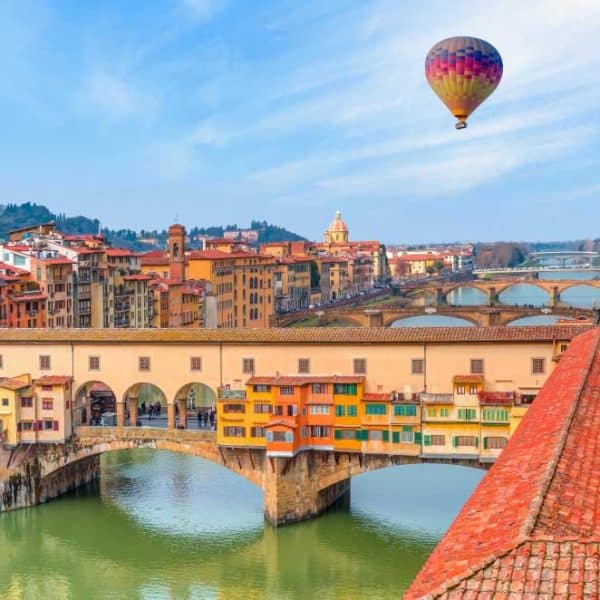 Vista panorâmica de Florença com a Ponte Vecchio sobre o rio Arno, prédios coloridos de telhados vermelhos e um balão de ar quente flutuando no céu azul.