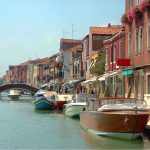 Canal estreito com água azul-esverdeada, barcos atracados nas laterais e casas coloridas com varandas e flores; ao fundo, uma pequena ponte de pedra sob céu claro.