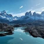 Paisagem da Patagônia com montanhas nevadas, lago de águas azul-turquesa e formações rochosas sob céu azul, cenário natural intocado e panorâmico.