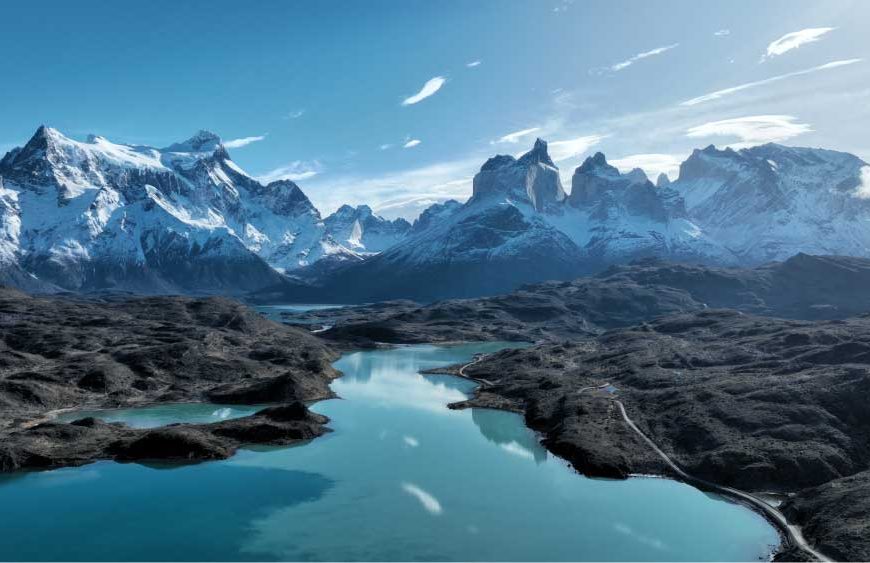Paisagem da Patagônia com montanhas nevadas, lago de águas azul-turquesa e formações rochosas sob céu azul, cenário natural intocado e panorâmico.