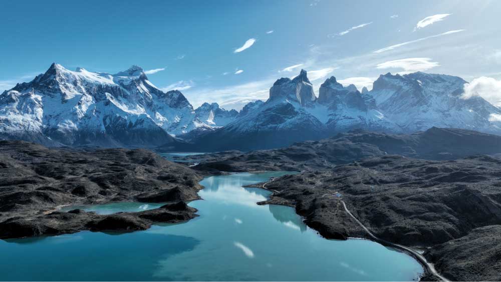Paisagem da Patagônia com montanhas nevadas, lago de águas azul-turquesa e formações rochosas sob céu azul, cenário natural intocado e panorâmico.