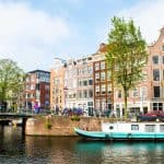 Canal de Amsterdã com casas históricas estreitas e coloridas ao fundo, uma ponte com pedestres e bicicletas, árvores verdes nas margens e um barco atracado na água em um dia ensolarado