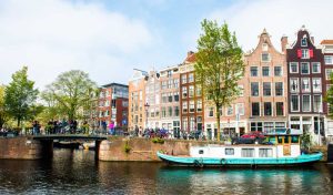 Canal de Amsterdã com casas históricas estreitas e coloridas ao fundo, uma ponte com pedestres e bicicletas, árvores verdes nas margens e um barco atracado na água em um dia ensolarado
