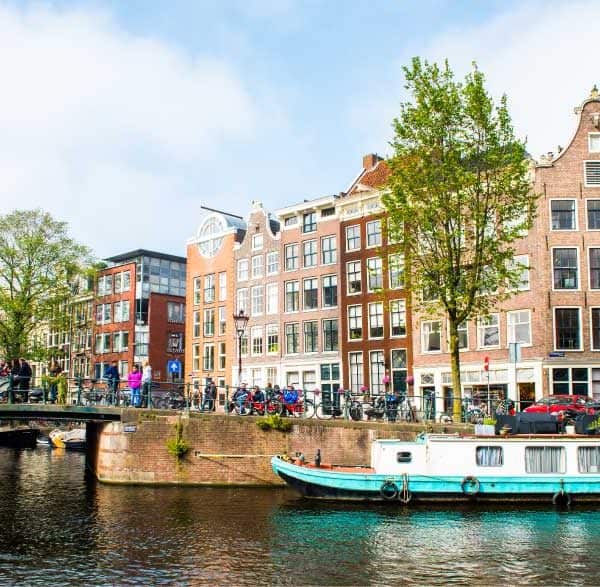 Canal de Amsterdã com casas históricas estreitas e coloridas ao fundo, uma ponte com pedestres e bicicletas, árvores verdes nas margens e um barco atracado na água em um dia ensolarado