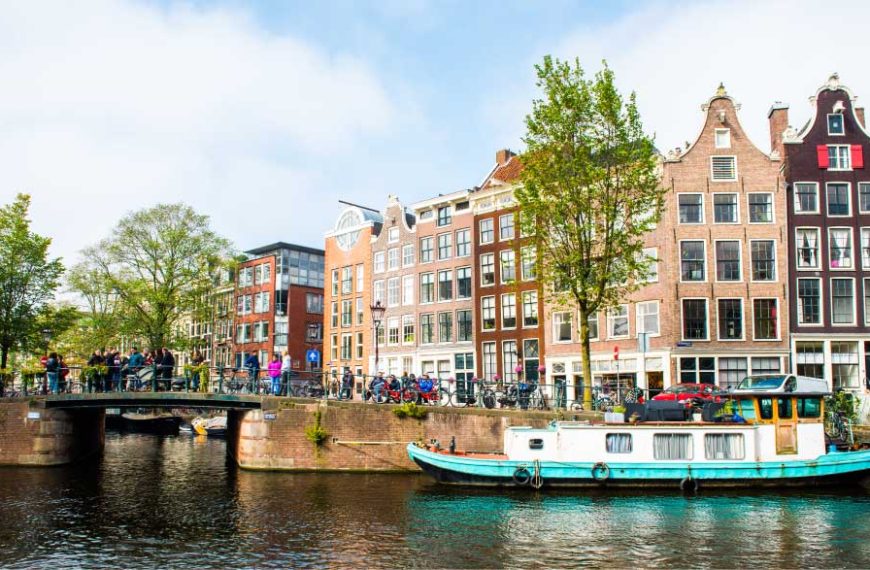 Canal de Amsterdã com casas históricas estreitas e coloridas ao fundo, uma ponte com pedestres e bicicletas, árvores verdes nas margens e um barco atracado na água em um dia ensolarado