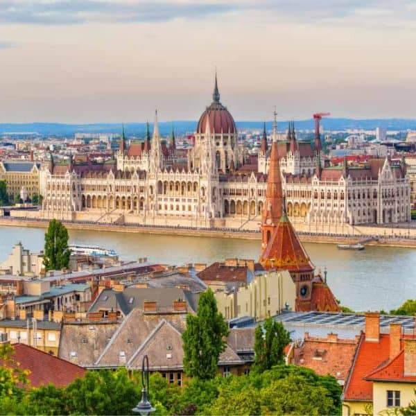 Vista panorâmica do Parlamento de Budapeste às margens do rio Danúbio, com arquitetura histórica da Hungria, telhados da cidade em primeiro plano e céu azul ao fundo.