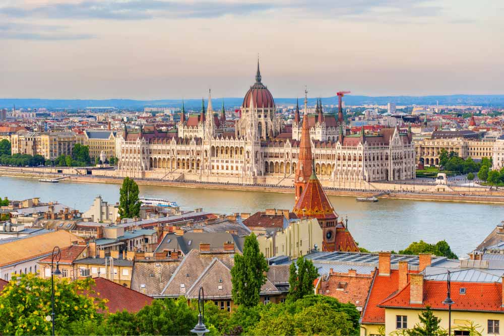 Vista panorâmica do Parlamento de Budapeste às margens do rio Danúbio, com arquitetura histórica da Hungria, telhados da cidade em primeiro plano e céu azul ao fundo.
