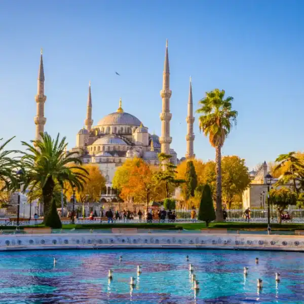 Mesquita Azul (Sultan Ahmed Mosque) em Istambul, com seus seis minaretes e grande cúpula, vista ao fundo de um jardim com palmeiras, árvores outonais e um espelho d’água sob céu azul.