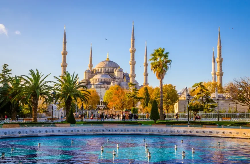 Mesquita Azul (Sultan Ahmed Mosque) em Istambul, com seus seis minaretes e grande cúpula, vista ao fundo de um jardim com palmeiras, árvores outonais e um espelho d’água sob céu azul.