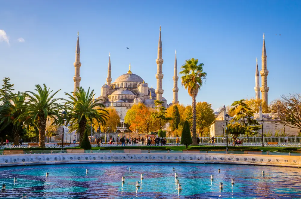 Mesquita Azul (Sultan Ahmed Mosque) em Istambul, com seus seis minaretes e grande cúpula, vista ao fundo de um jardim com palmeiras, árvores outonais e um espelho d’água sob céu azul.