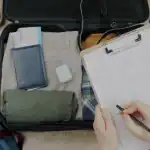 Pessoa organizando itens de viagem em uma mala, checklist de viagem ao lado.