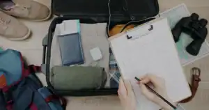 Pessoa organizando itens de viagem em uma mala, checklist de viagem ao lado.