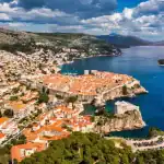 Vista aérea da cidade histórica de Dubrovnik, na Croácia, com muralhas medievais, telhados de cerâmica laranja e o Mar Adriático azul ao fundo, cercada por montanhas e céu parcialmente nublado.