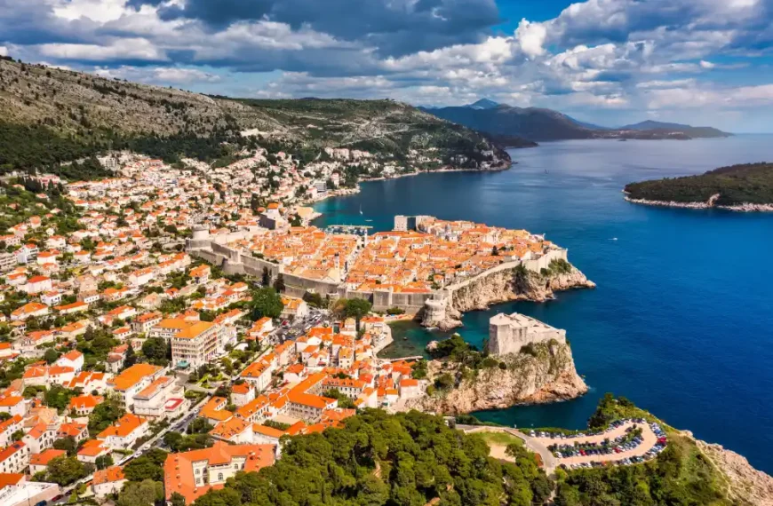 Vista aérea da cidade histórica de Dubrovnik, na Croácia, com muralhas medievais, telhados de cerâmica laranja e o Mar Adriático azul ao fundo, cercada por montanhas e céu parcialmente nublado.