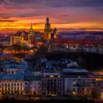 Vista panorâmica do Castelo de Wawel, em Cracóvia, na Polônia, iluminado ao entardecer, com céu alaranjado e roxo ao fundo e prédios históricos da cidade em primeiro plano