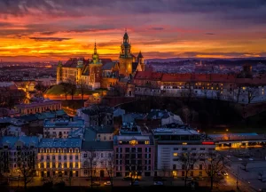 Vista panorâmica do Castelo de Wawel, em Cracóvia, na Polônia, iluminado ao entardecer, com céu alaranjado e roxo ao fundo e prédios históricos da cidade em primeiro plano