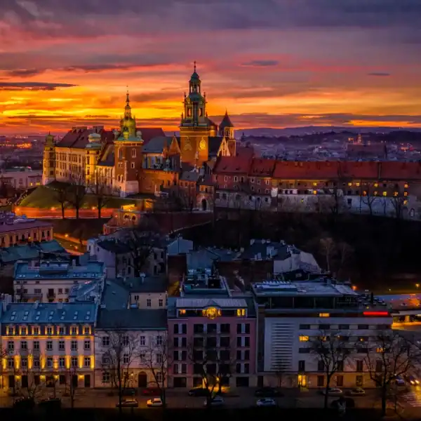 Vista panorâmica do Castelo de Wawel, em Cracóvia, na Polônia, iluminado ao entardecer, com céu alaranjado e roxo ao fundo e prédios históricos da cidade em primeiro plano
