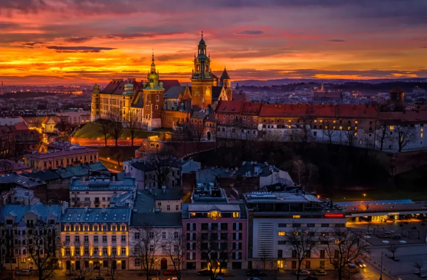 Vista panorâmica do Castelo de Wawel, em Cracóvia, na Polônia, iluminado ao entardecer, com céu alaranjado e roxo ao fundo e prédios históricos da cidade em primeiro plano