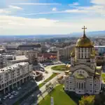 Vista aérea de uma cidade com uma grande igreja de cúpula dourada em primeiro plano, cercada por ruas, prédios residenciais e áreas verdes. Ao fundo, a paisagem se estende até colinas sob um céu azul com nuvens leves.