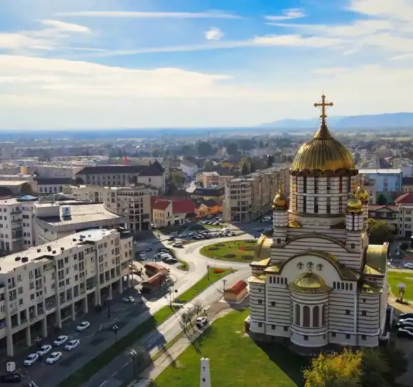 Vista aérea de uma cidade com uma grande igreja de cúpula dourada em primeiro plano, cercada por ruas, prédios residenciais e áreas verdes. Ao fundo, a paisagem se estende até colinas sob um céu azul com nuvens leves.