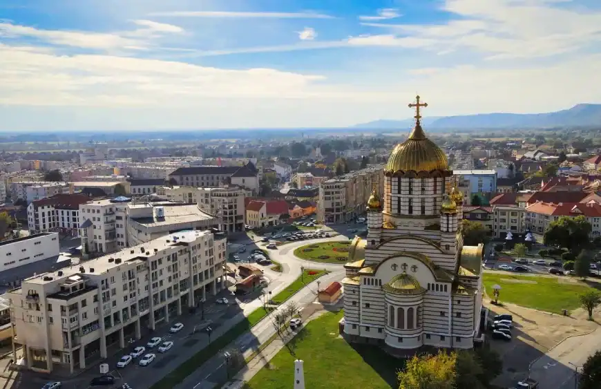 Vista aérea de uma cidade com uma grande igreja de cúpula dourada em primeiro plano, cercada por ruas, prédios residenciais e áreas verdes. Ao fundo, a paisagem se estende até colinas sob um céu azul com nuvens leves.