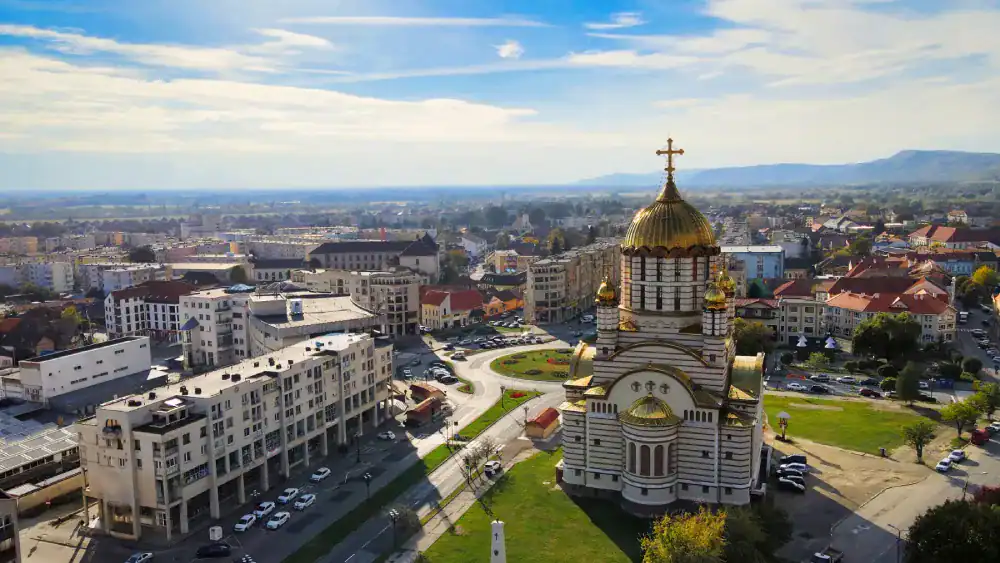 Vista aérea de uma cidade com uma grande igreja de cúpula dourada em primeiro plano, cercada por ruas, prédios residenciais e áreas verdes. Ao fundo, a paisagem se estende até colinas sob um céu azul com nuvens leves.