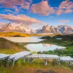 Mirante de madeira com vista para lagoas e montanhas nevadas no Torres del Paine National Park, sob céu azul com nuvens ao pôr do sol.