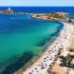 Vista aérea de uma praia de águas cristalinas em tons de azul-turquesa e verde, com faixa de areia cheia de banhistas e guarda-sóis. Ao fundo, pequenas ilhas rochosas conectadas por uma estreita faixa de terra e o mar azul intenso se estendendo até o horizonte sob céu limpo.