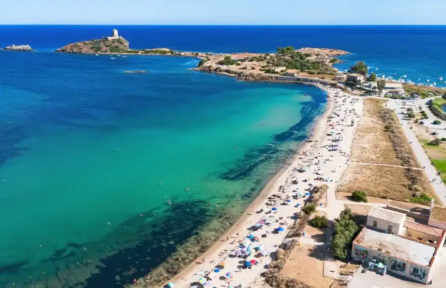 Vista aérea de uma praia de águas cristalinas em tons de azul-turquesa e verde, com faixa de areia cheia de banhistas e guarda-sóis. Ao fundo, pequenas ilhas rochosas conectadas por uma estreita faixa de terra e o mar azul intenso se estendendo até o horizonte sob céu limpo.