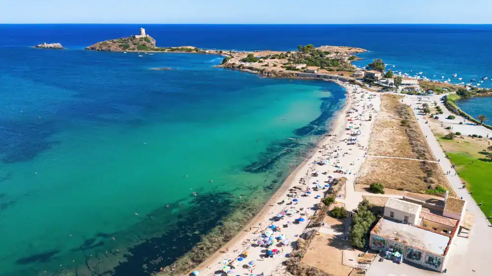 Vista aérea de uma praia de águas cristalinas em tons de azul-turquesa e verde, com faixa de areia cheia de banhistas e guarda-sóis. Ao fundo, pequenas ilhas rochosas conectadas por uma estreita faixa de terra e o mar azul intenso se estendendo até o horizonte sob céu limpo.