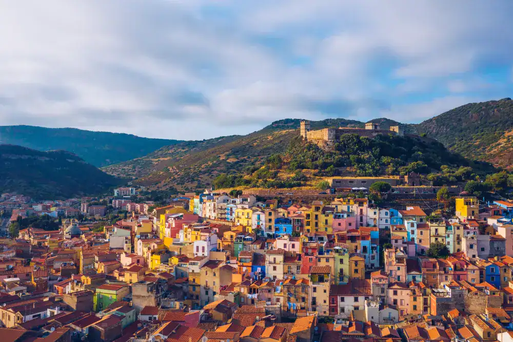 Vista panorâmica de uma cidade histórica com casas coloridas e telhados em tons terrosos espalhados pelas colinas. No alto de uma montanha ao fundo, uma fortaleza antiga domina a paisagem cercada por vegetação, sob céu parcialmente nublado.
