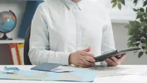 Imagem de uma pessoa segurando um tablet, com mapas e documentos de viagem sobre a mesa, simbolizand.