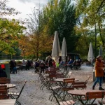Café ao ar livre à beira de um lago, com mesas, cadeiras e pessoas aproveitando um dia de outono entre árvores.