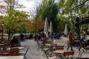 Café ao ar livre à beira de um lago, com mesas, cadeiras e pessoas aproveitando um dia de outono entre árvores.