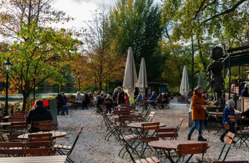 Café ao ar livre à beira de um lago, com mesas, cadeiras e pessoas aproveitando um dia de outono entre árvores.