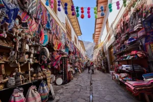 Rua estreita de pedra em um mercado tradicional andino, cercada por lojas repletas de artesanato colorido, tecidos, bolsas, chapéus e lembranças típicas. Pompons multicoloridos estão pendurados sobre a via, enquanto visitantes caminham pelo corredor com montanhas ao fundo sob céu azul.